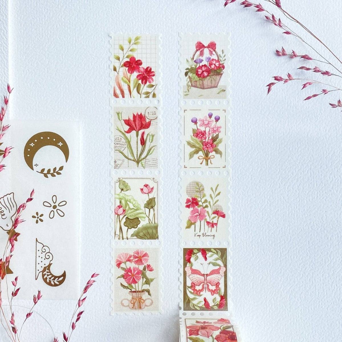 A set of floral washi tapes arranged on a textured white background, alongside a sheet of decorative washi tapes featuring a gold crescent moon and floral motifs, accented by dried pink flowers in the corners.