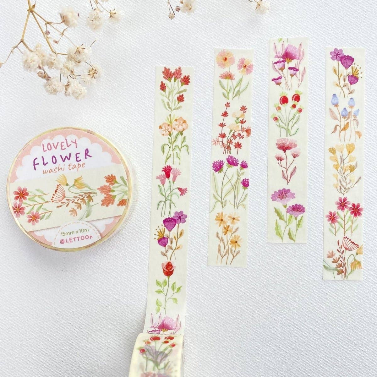 A roll of floral washi tape and four narrow strips displayed on a light textured background, with dried white flowers as props. The tape features watercolor-style wildflowers in pink, red, purple, yellow, and green on an off-white base.