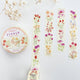 A roll of floral washi tape and four narrow strips displayed on a light textured background, with dried white flowers as props. The tape features watercolor-style wildflowers in pink, red, purple, yellow, and green on an off-white base.