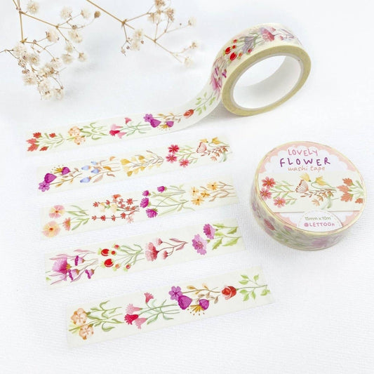 A roll of floral washi tape and four narrow strips displayed on a light textured background, with dried white flowers as props. The tape features watercolor-style wildflowers in pink, red, purple, yellow, and green on an off-white base.