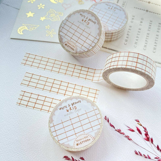 Three rolls of washi tape with a white and brown grid pattern are arranged on a textured white surface alongside matching strips and decorative gold foil paper. The labels on the rolls read 'WHITE & BROWN GRID washi tape' and show the brand 'Lettoo'.