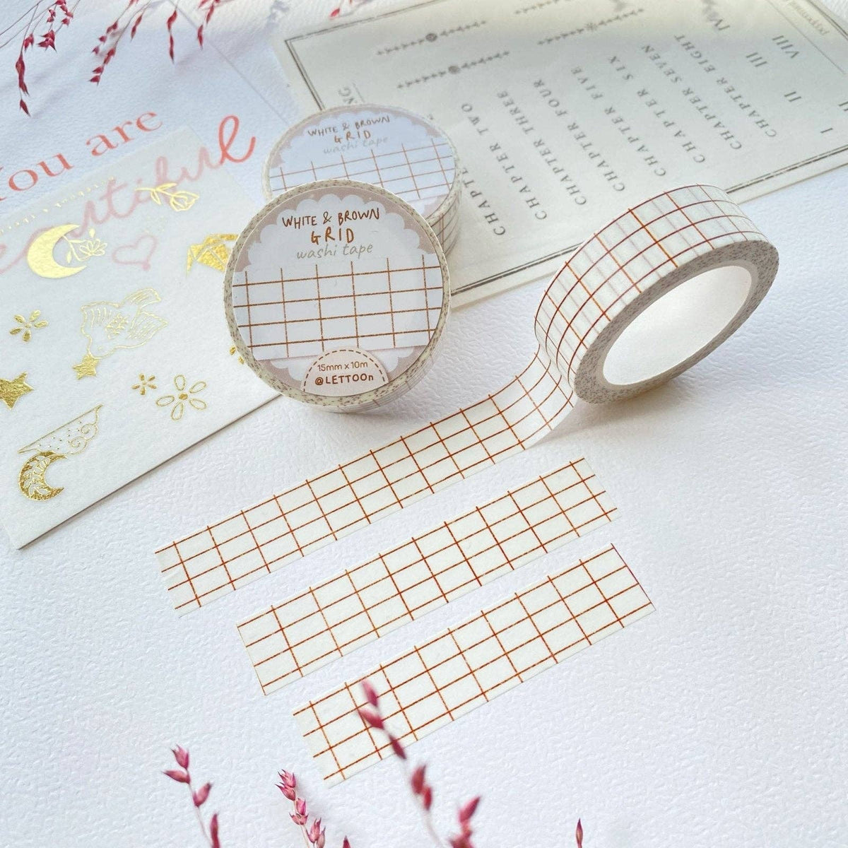 Three rolls of washi tape with a white base and a thin orange-brown grid pattern, displayed on a white textured surface with decorative stationery props.