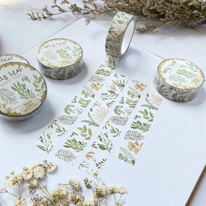 Three rolls of washi tape with a botanical leaf pattern are arranged on a white textured surface with dried flowers. The tape features green, beige, and brown leaves on a white background.