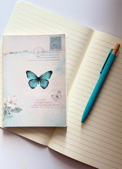 A decorative notebook with a vintage-style cover featuring a blue butterfly and floral illustration, placed on top of an open lined notebook with a teal pen beside it.
