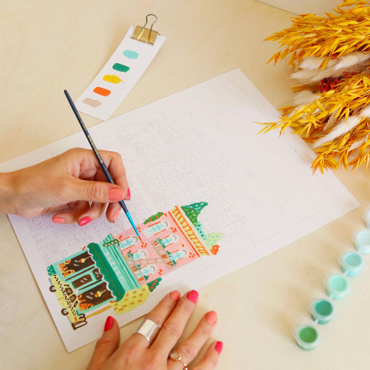 Person painting a colorful illustration on paper with watercolors, surrounded by paint tubes and a color chart.