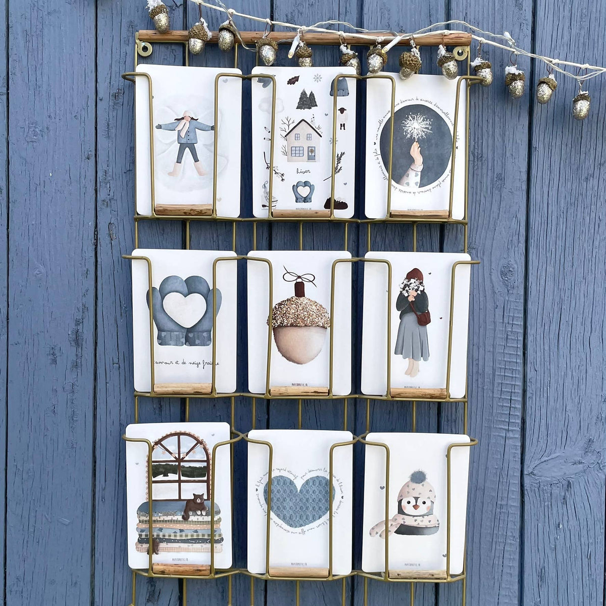 A wall-mounted gold metal grid organizer holding nine white fabric pouches or small bags, each printed with a different winter-themed illustration such as a snowman, acorn, heart, and snowflake. The organizer is hung against a blue wooden plank wall with decorative string lights above.