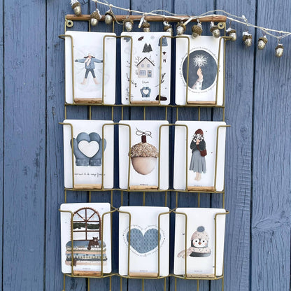 A wall-mounted gold metal grid organizer holding nine white fabric pouches or small bags, each printed with a different winter-themed illustration such as a snowman, acorn, heart, and snowflake. The organizer is hung against a blue wooden plank wall with decorative string lights above.