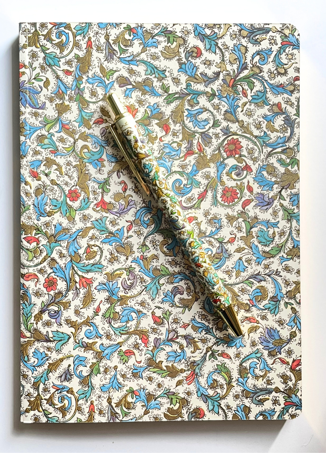 A decorative notebook with an intricate floral and scroll pattern in blue, red, green, and gold on a cream background, with a gold pen resting diagonally across the top.