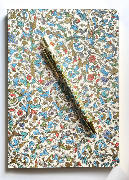 A decorative notebook with an intricate floral and scroll pattern in blue, red, green, and gold on a cream background, with a gold pen resting diagonally across the top.
