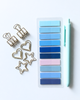 A flat-lay of stationery items on a light gray background: a clear plastic sheet with rectangular sticky notes in shades of blue, navy, and pale pink, next to a mint green pen and gold binder clips shaped like hearts and stars.