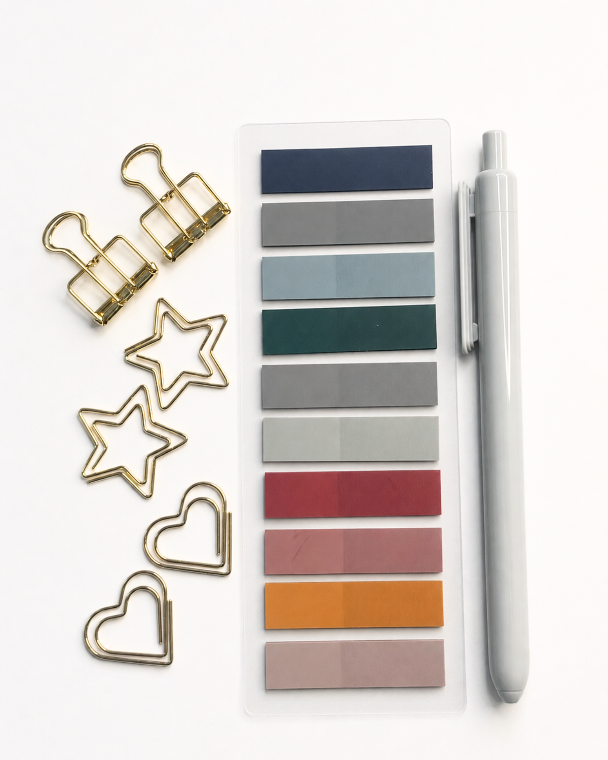 A flat-lay of stationery accessories on a white background, including gold metal binder clips, star and heart clips, a clear plastic swatch card with multiple color tabs, and a light gray pen.