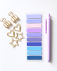 A flat-lay of stationery items on a light gray background: a lavender pen, a sheet of rectangular sticky notes in pastel shades from light purple to blue, and gold metal paperclips in binder, heart, and star shapes.