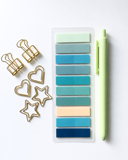 A set of pastel and teal sticky notes in a clear plastic sleeve, styled with gold paperclips, heart and star clipboards, and a light green pen on a light gray background.