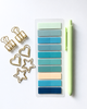 A set of pastel and teal sticky notes in a clear plastic sleeve, styled with gold paperclips, heart and star clipboards, and a light green pen on a light gray background.