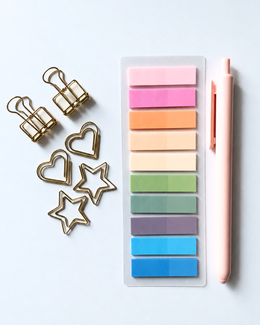 A flat-lay of stationery items on a light gray surface: a clear plastic holder filled with pastel sticky notes in pink, purple, orange, beige, green, teal, lavender, and blue, next to a pale pink pen and gold metal paperclips shaped like hearts and stars.