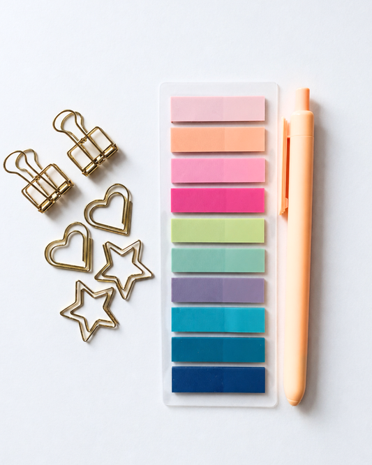 A set of pastel sticky notes in a clear plastic sleeve next to a peach pen, with gold metal binder clips and star, heart, and oval clips arranged on a white background.