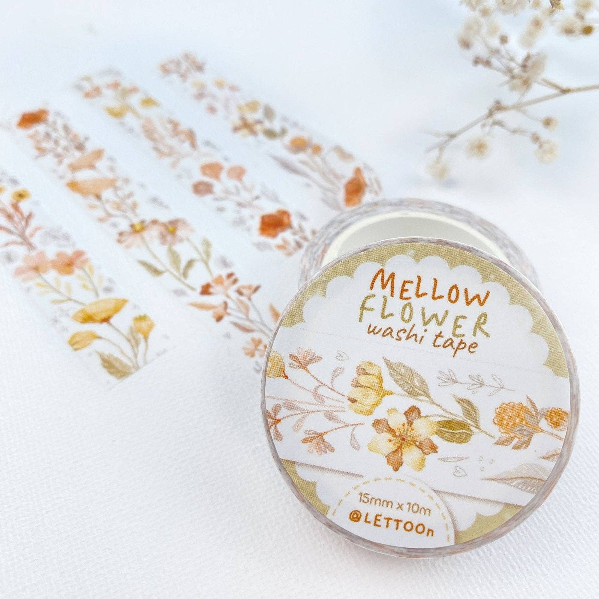 A roll of washi tape labeled 'Mellow Flower washi tape' by LETTOON, shown with several strips unrolled on a soft white fabric background with dried flowers as props.