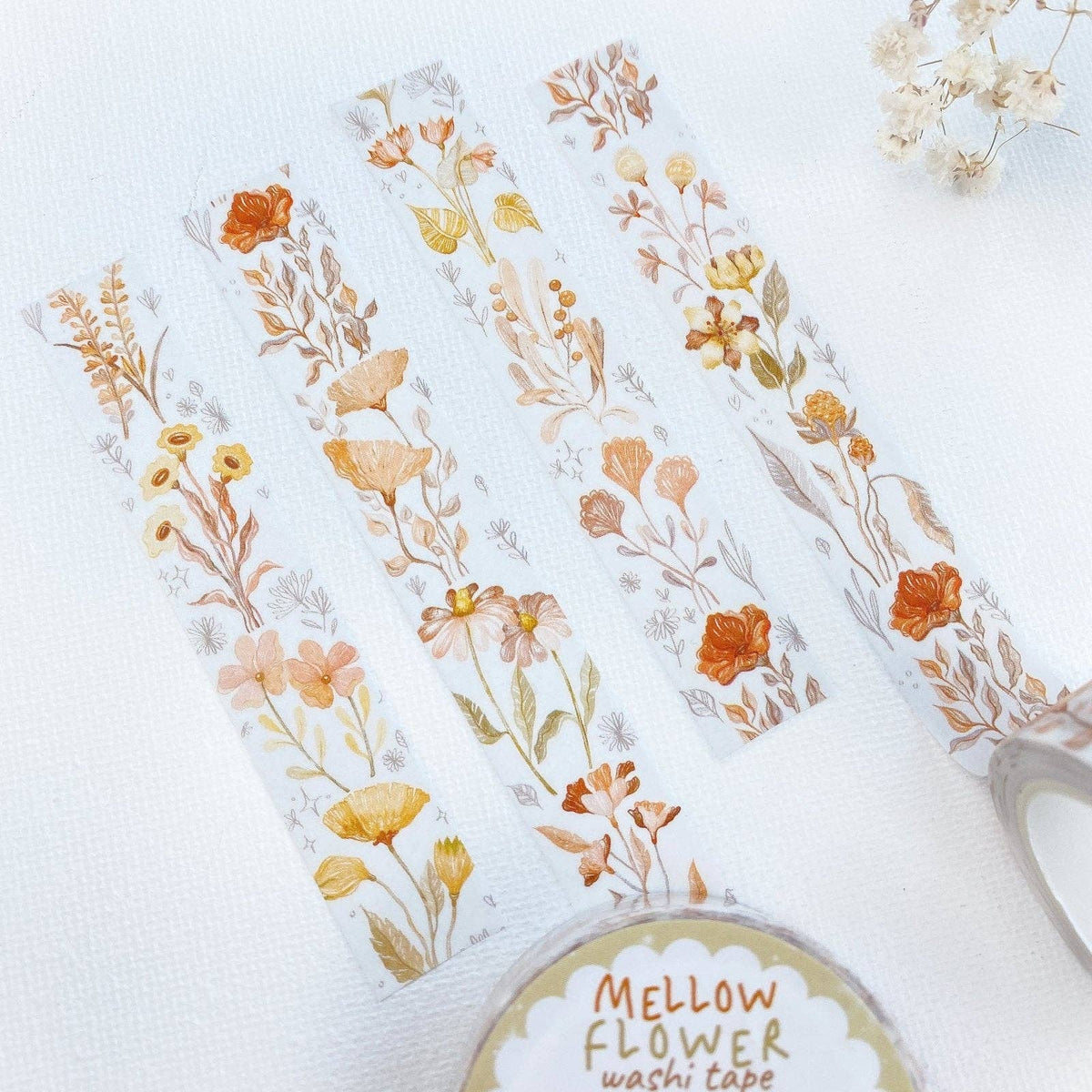 Four strips of washi tape with delicate watercolor-style wildflower designs in soft orange, yellow, beige, and muted green on a white background. A roll of Mellow Flower washi tape is partially visible at the bottom right, along with a small dried flower sprig in the top right corner.