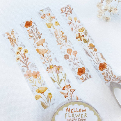 Four strips of washi tape with delicate watercolor-style wildflower designs in soft orange, yellow, beige, and muted green on a white background. A roll of Mellow Flower washi tape is partially visible at the bottom right, along with a small dried flower sprig in the top right corner.