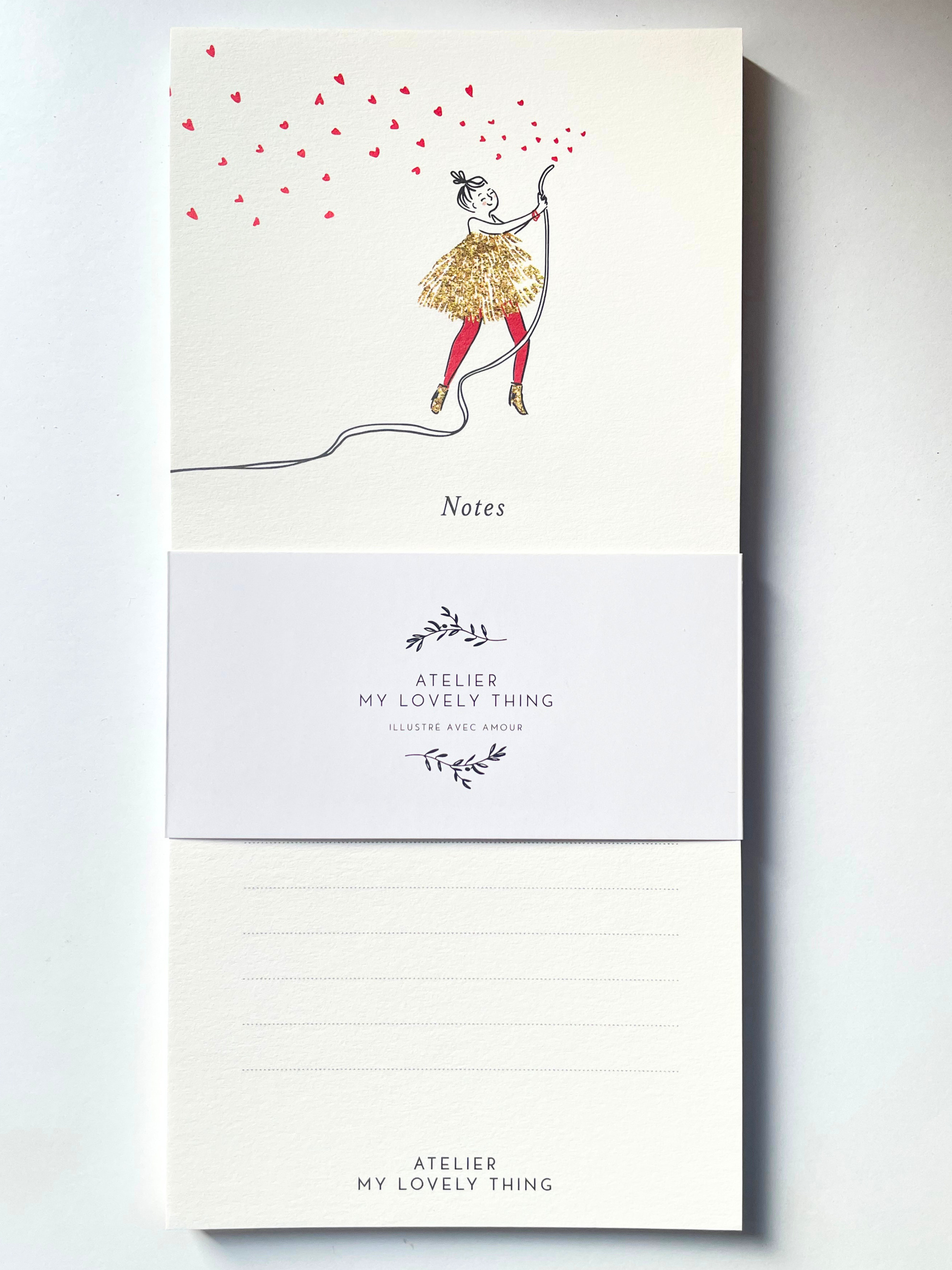A vertical cream-colored notepad with a whimsical illustration of a girl in a glittery gold dress and red tights, holding a string that casts many red heart-shaped shadows. The notepad is wrapped in a white paper band printed with the brand name 'ATELIER MY LOVELY THING' and features lined writing space on the back.