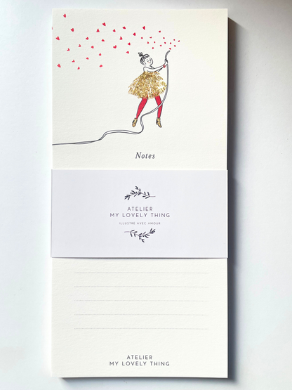 A vertical cream-colored notepad with a whimsical illustration of a girl in a glittery gold dress and red tights, holding a string that casts many red heart-shaped shadows. The notepad is wrapped in a white paper band printed with the brand name 'ATELIER MY LOVELY THING' and features lined writing space on the back.