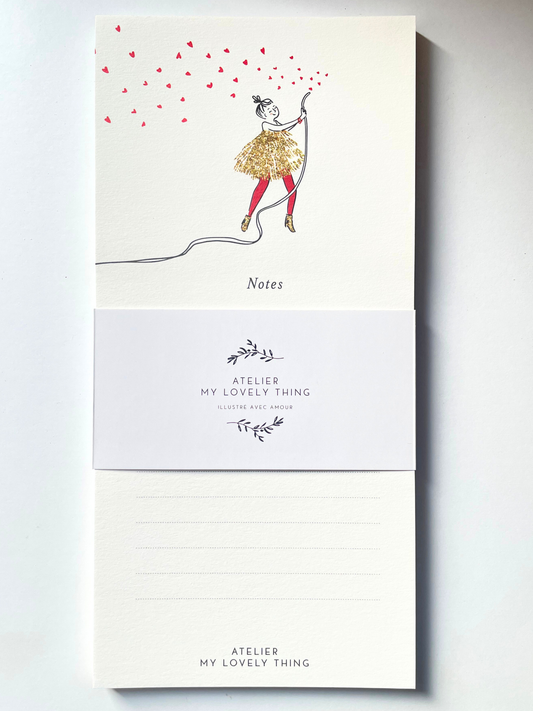 A vertical cream-colored notepad with a whimsical illustration of a girl in a glittery gold dress and red tights, holding a string that casts many red heart-shaped shadows. The notepad is wrapped in a white paper band printed with the brand name 'ATELIER MY LOVELY THING' and features lined writing space on the back.