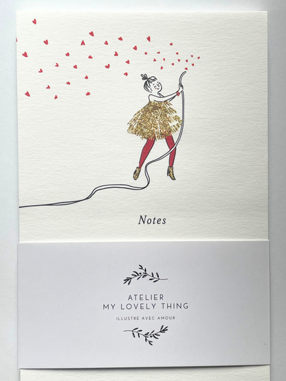 A white textured card or notebook cover with a whimsical illustration of a girl in a gold tutu and red tights jumping while pulling a long black string, with small red heart shapes floating above. The word 'Notes' appears below the illustration.