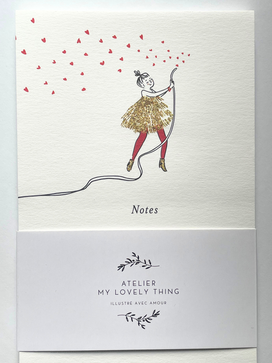 A vertical cream-colored notepad with a whimsical illustration of a girl in a glittery gold dress and red tights, holding a string that casts many red heart-shaped shadows. The notepad is wrapped in a white paper band printed with the brand name 'ATELIER MY LOVELY THING' and features lined writing space on the back.