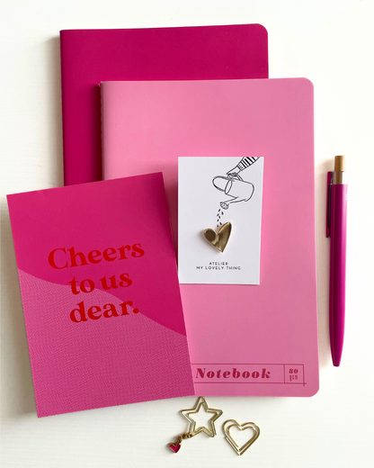 Pink notebook with 'Cheers to us dear' text, a pink pen, and decorative heart and star stickers on a white background.