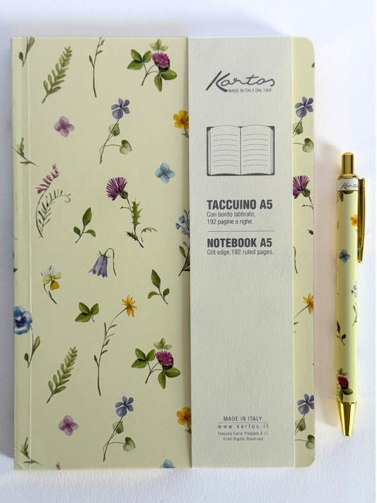 A Kartos Taccuino A5 notebook with a cream-colored cover decorated with delicate watercolor wildflowers, shown next to a matching floral gold and white pen on a clean white background.