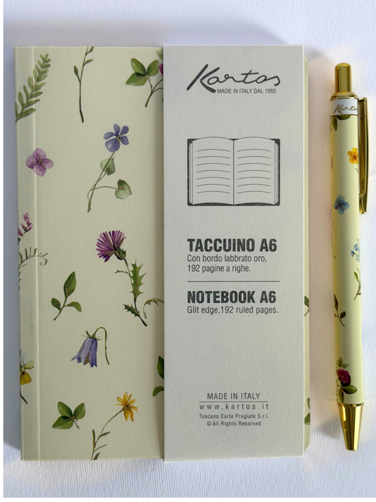 A Kartos Taccuino A6 notebook with a cream-colored cover decorated with delicate botanical illustrations, shown next to its matching pen and a vertical product information card.