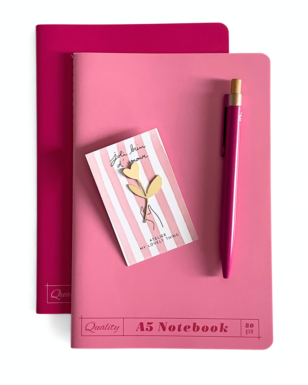 A pink A5 notebook with a matching pink pen and a small striped notepad, arranged on top of a darker pink notebook against a white background.