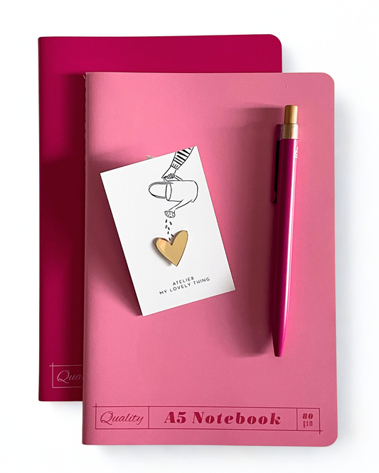 A pink A5 notebook with a matching pink pen and a small gold heart pin, arranged on top of a darker pink notebook against a clean white background.